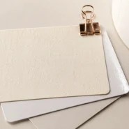 Bagasse label stock swatch, blank and unprinted, shown with gloss and matte swatches and a partial label roll on a warm off-white surface.